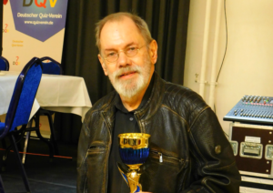 Klaus Otto Nagorsnik mit einem Siegerpokal in den Händen vor dem Wappen des Deutschen Quizvereins